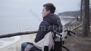 沮丧的白人男孩坐在轮椅上独自在河岸度过秋日。 可怜的残疾青年画像